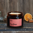 Load image into Gallery viewer, A lit Christmas scented candle with a label that reads 'Christmas', alongside cinnamon sticks and a slice of orange, against a wooden background.