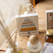 Load image into Gallery viewer, A square card or matchbox-style product standing on a wooden table, printed with an illustration of a tiered birthday cake and the words 'Birthday Matches.' The product is photographed in a styled setting with blurred bottles and decor in the background.

