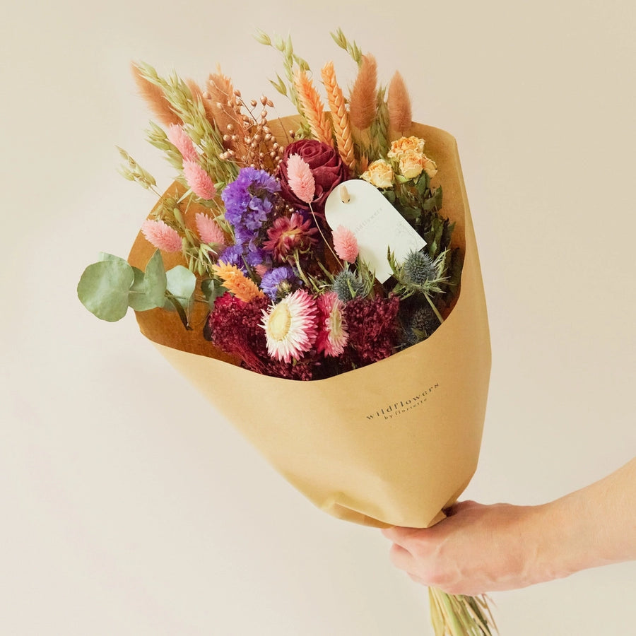 Close up of a meium bouquet of burgundy wild flowers on a white background showing plants such as eucalptyus, Rose, thistles and dried grasses. 