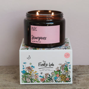 Stargazer Flora Scented Candle. Our candles are all hand poured with natural plant based wax. Hand poured in the UK and poured in a brown amber glass by our female owned team. Long lasting candles that fully scent a room. Our candles are complete with a gold alliminium lid and then packaged in a beautiful botanical box, which features candle safety instructions in a cottage core theme. They are long lasting candles, rated excellent on Trust Pilot and fully scent a room. Sheer Lily White rose and Jasmine. 