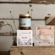 Load image into Gallery viewer, An amber glass jar candle labeled 'Sea Salt' by The Flora Lab, stacked on top of its illustrated white box, next to a matching 'The Swimmer' luxury matches box, styled on a rustic wooden surface against a whitewashed wood background.
