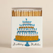 Load image into Gallery viewer, A boxed set of birthday matches with a white matchbox cover printed with an illustration of a tiered birthday cake on a blue stand and the words 'Birthday Matches.' The matches inside are light wood with white tips, and the box is shown open against a plain warm beige background.
