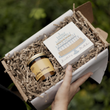 Load image into Gallery viewer, A gift box filled with shredded brown paper crinkle paper, containing a small jar candle with a gold label and a square card with a birthday cake illustration and the words 'Birthday matches'. A hand is holding the open box against a blurred green outdoor background.
