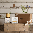 Load image into Gallery viewer, A curated gift set from The Flora Lab displayed on a rustic wooden crate. The set includes a candle in a glass jar with a gold lid, a small floral box, a square birthday card with a birthday cake illustration, and a large brown cardboard gift box tied with a cream ribbon.
