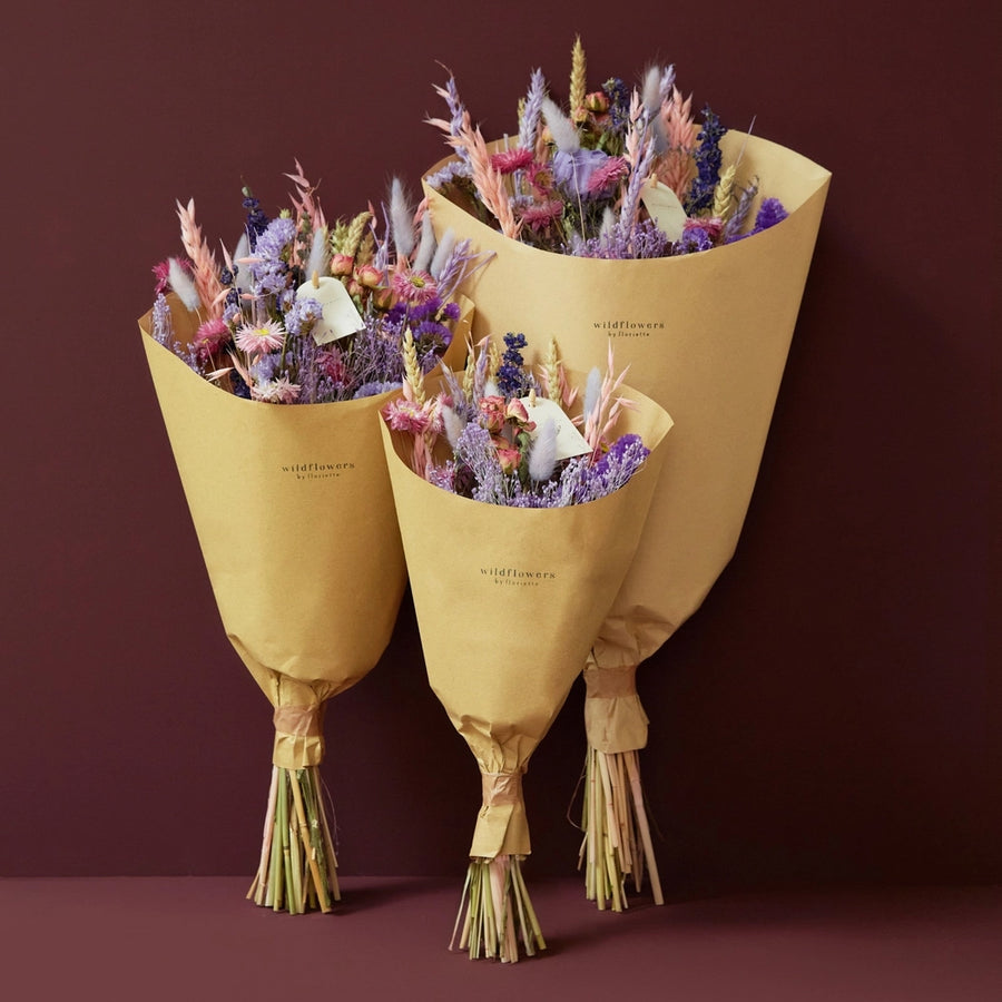 Purple Glow Dried Wild flower bouquet. 
Illuminate your space with the enchanting hues of the Purple Glow bouquet from our Autumn Collection. This hand-tied field bouquet combines soft pinks, lilacs, blues, and purples for a magical and elegant display.

This bouquet contains: Light pink oats, natural wheat, lilac wheat, blue delphinium, Odillia spray roses, lavender statice, blue statice, purple limonium, pink acroclinium/rhodanthe, milka shola, and milka broom bloom.