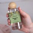 Load image into Gallery viewer, A close-up of a hand holding The Flora Lab Renaissance Man Room Perfume Spray, a clear glass bottle with a botanical label noting scent notes of orange, bergamot and grapefruit, topped with a wooden ball lid
