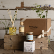 Load image into Gallery viewer, A curated gift set from The Flora Lab featuring a brown cardboard box with a cream ribbon, a reed diffuser bottle with wooden sticks, a small glass candle jar, and a Sea Salt scented box, arranged on rustic wooden crates against a weathered white wood wall.
