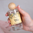Load image into Gallery viewer, Hand holding a clear glass perfume bottle with a wooden cap and floral label.
