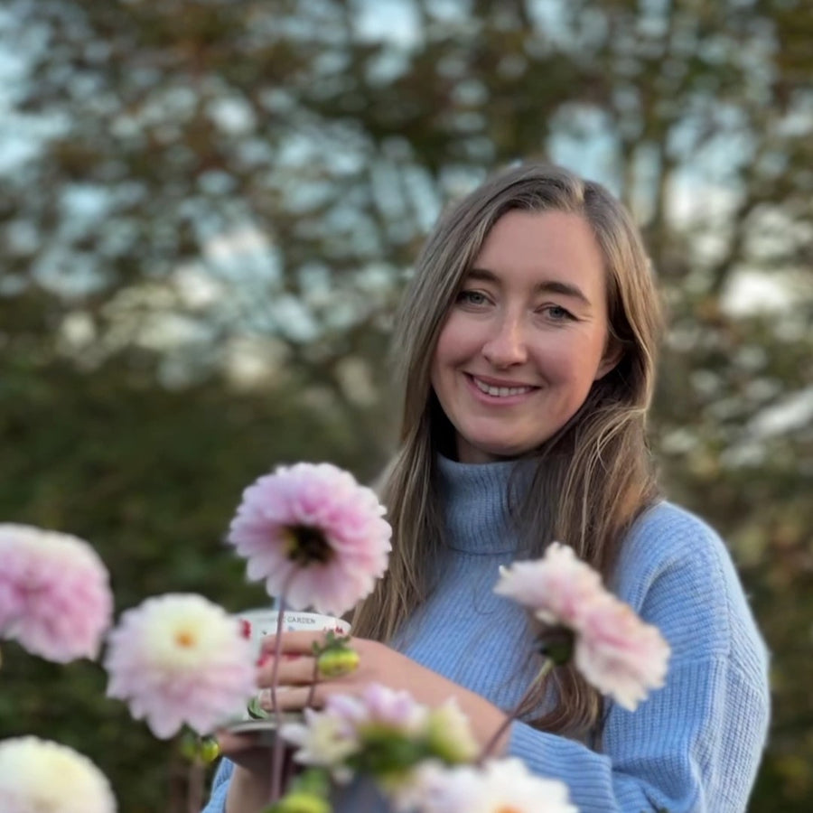 Image of a female founder in flowers and nature, celebrating a business that is inspired by nature. the flowers that feature are pink dahlia and the brand owner is holding a cup of tea. 