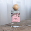 Load image into Gallery viewer, The Flora Lab Stargazer Hand Poured Room Perfume Spray in a clear glass bottle with a wooden ball lid, featuring a pink floral label, resting on a rustic wooden surface

