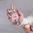 Load image into Gallery viewer, A close-up of a hand holding the The Flora Lab Stargazer Room Perfume Spray featuring sheer lily white rose and jasmine feminine scent , a compact clear glass bottle with a pink floral label noting scent notes of lily, ylang ylang and white rose, topped with a wooden ball lid
