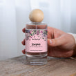 Load image into Gallery viewer, A hand holding The Flora Lab Stargazer Room Perfume Spray, a clear glass bottle with a pink botanical label and wooden ball lid, placed on a wooden surface against a soft white background
