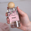 Load image into Gallery viewer, A close-up of a hand holding the The Flora Lab Stargazer Room Perfume Spray, a compact clear glass bottle with a pink floral label noting scent notes of lily, ylang ylang and white rose, topped with a wooden ball lid
