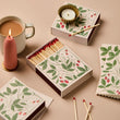 Load image into Gallery viewer, A styled flat-lay of decorative matchboxes with a botanical strawberry print, including one open box of wooden matches with red tips and a small gold cup holder insert, alongside a pink candle, a mug of coffee, and scattered matchsticks on a warm beige background.
