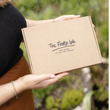 Load image into Gallery viewer, A person holding a brown kraft paper box printed with the brand name 'The Flora Lab' and the text 'Hand Poured Natural Wax Candles' against a blurred green outdoor background.
