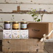 Load image into Gallery viewer, Three jars of The Flora Lab candles displayed on top of their illustrated boxes, next to a large brown cardboard gift box tied with a cream ribbon, all arranged on a rustic wooden crate against a whitewashed wood background with decorative greenery.
