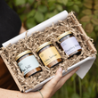 Load image into Gallery viewer, A gift box containing three small jars of probiotic supplements nestled in brown paper shred, held by a person outdoors with green foliage in the background.
