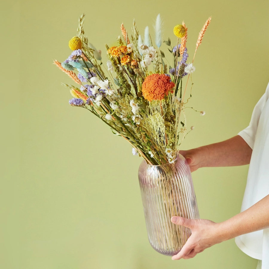 Image of pastel dried wildflower bouquet in a see through vase on a green background.  All sustainably grown and dried. This bouquet contains: Natural oats, natural wheat, pesca wheat, light blue phalaris, light blue lagurus, craspedia, trinity spray roses, green amaranthus, lavender statice, white rhodanthe, and frosted salmon achillea.